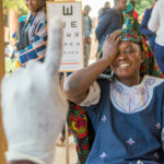 A woman with a white, red and blue dress is sitting on a chair, raising her right hand on top of her right eye whilst smiling. Next to her there is a vision test with letters. Right in front of her there is a blurry hand wearing a white glove and pointing to the ceiling with the index finger.