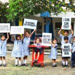 10 indian children outside holding advocacy signs for inclusion. Amongst them is a child sitting on a red rollable chair.