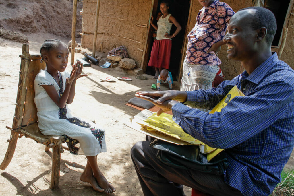 A man sits in front of a girl on the armchair and talks to her in sign language. She is wearing a white dress and looks at him intently while he explains with a smile.