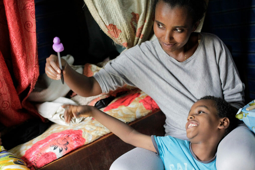 Abdi is sitting in his mother's lap. She is wearing a grey T-shirt and grey leggings, he is wearing a blue shirt. The boy smiles and points to a pen with a purple plastic duck that his mother is holding up.