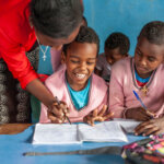Ein Kind mit Behinderung sitzt lachend neben seinen Klassenkameradinnen. Sie tragen alle einen rosa Pulli. Die Lehrerin beugt sich über das Kind und hält seine Hand, während es in dem Heft vor ihm zu schreiben versucht.