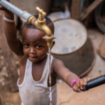 Small child looking at the camera while holding a water tank where water is coming out.