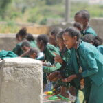 Girls from Ethiopia in green school uniforms stand in front of a well and wash their hands.