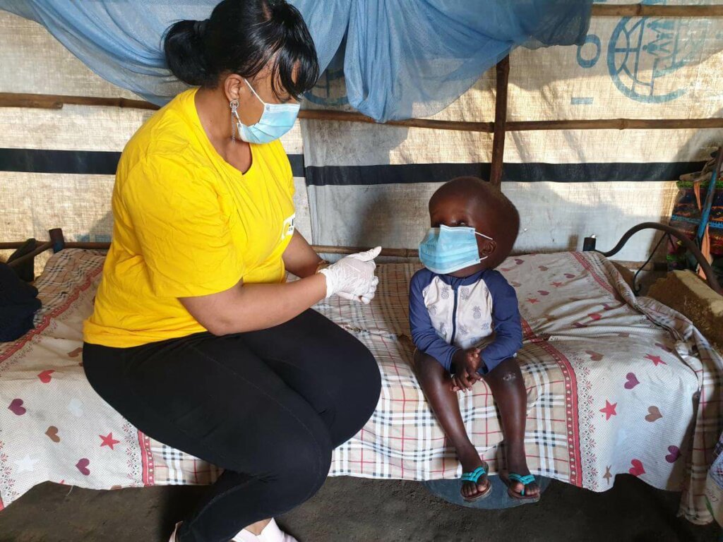 Sitting on a bed in a South Sudanese refugee camp Nyaush, a young girl with hydrocephalus talks to Light for the World country director Sophia Mohammed. Both wear protective masks.