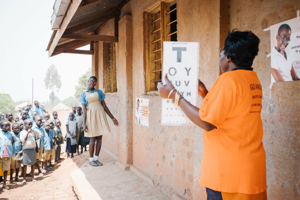 A eye health worker is holding up a vision screen test with letters. A girl student wearing glasses and the school uniform is a couple of meters in front of her leaning slightly to the right side with her head, trying to read the letters. Her school peers are behind her watching.