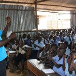 A southsudanese man stands in front of a class full of children talking to them in sign language.