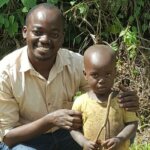 Father is kneeling down, holding his son next to him and both are looking into the camera.