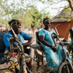 Two women in wheelchairs sit next to each other under a tree. Both are wearing blue clothes and look at each other amusedly.