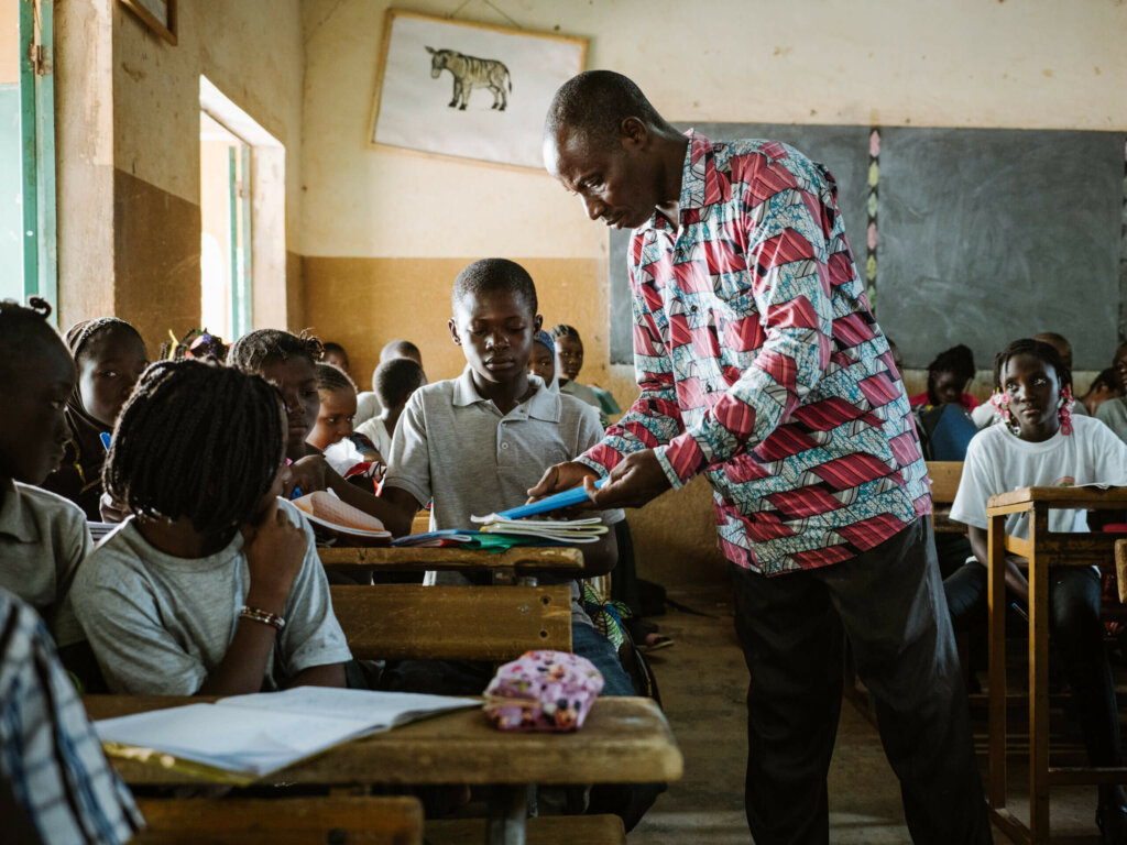 A teacher in a colourful shirt gives a boy back his notebook in the classroom.