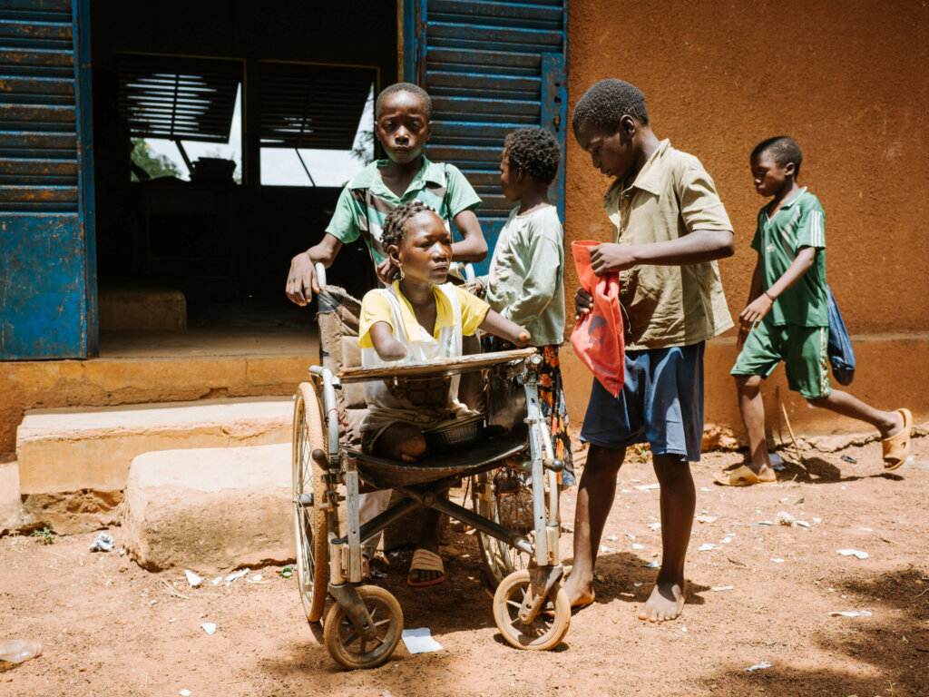 A young Burkinabe woman whose arms end at the ellbows sits in her wheelchair which is pushed by a younger boy. Three other youths are in the background.