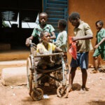 A young Burkinabe woman whose arms end at the ellbows sits in her wheelchair which is pushed by a younger boy. Three other youths are in the background.