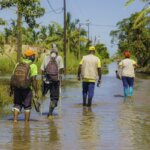 Four people, including Light for the World staff, walk through a muddy environment after another cyclone devastated Mozambique. All are wearing rubber boots and face masks.