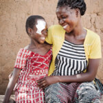 Young Burkinabe girl with her mother in Gaoua Burkina Faso. The girl wears a white eye bandage after her cataract surgery. Both are smiling.