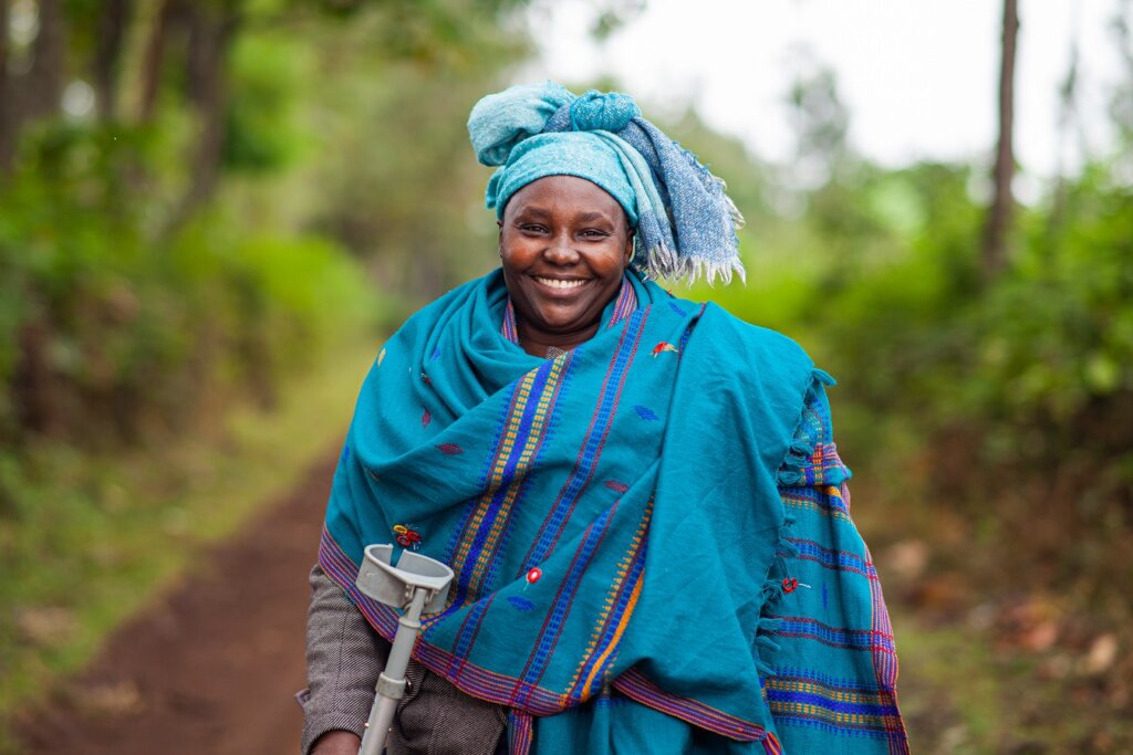 Woman wearing blue scarf and head scarf, and holding crutches, smiles into the camera. In the background is a rural road and trees.