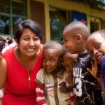 Nafisa Baboo smiling with a group of 6 Ethiopian children. Nafisa has lightbrown skin, short dark hear and wears a red sleaveless dress.