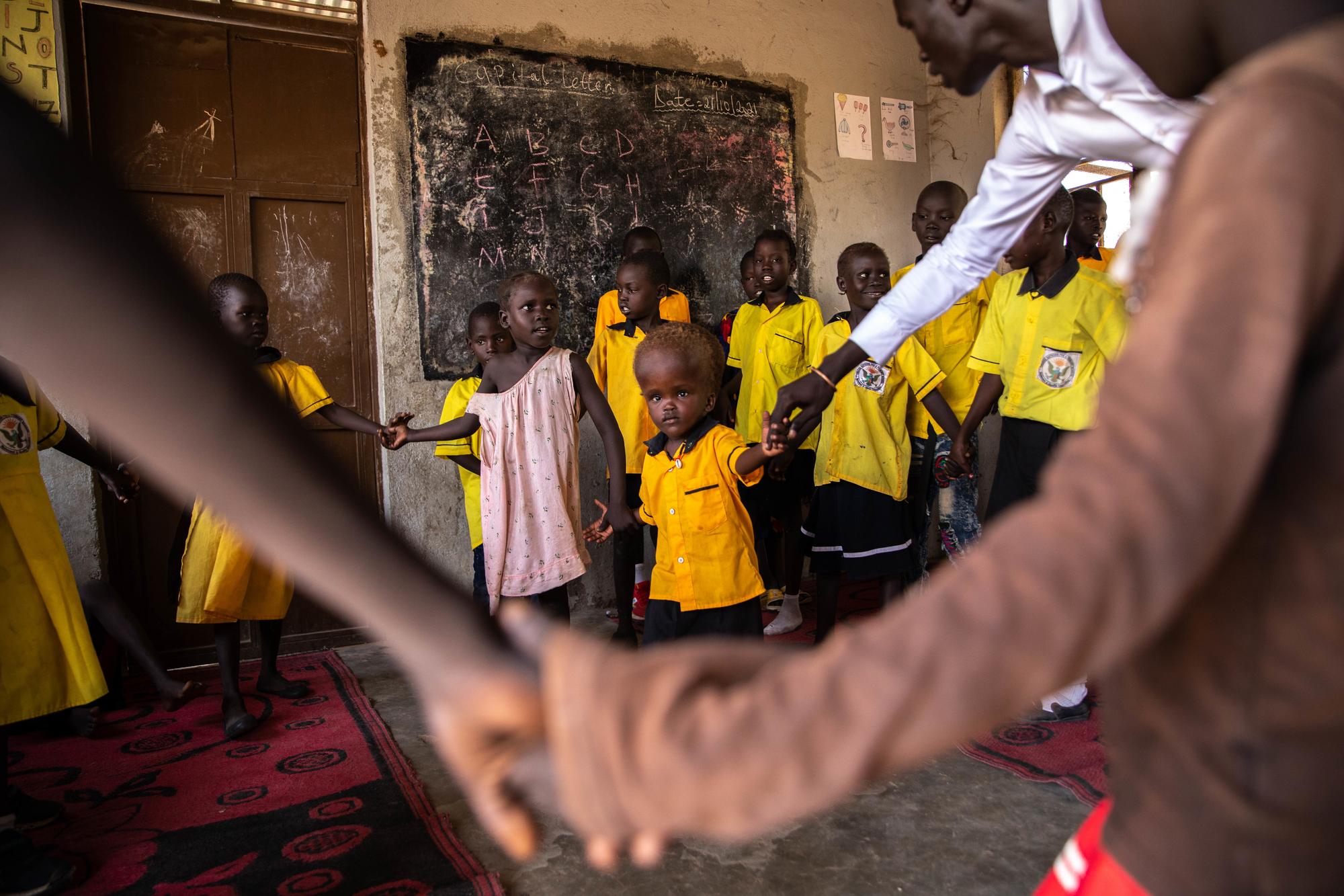 f-year old south sudanese girl nyamush has hydrocephalus. she is led by the hand of her teacher and is surrounded by her classmates, all wearing yellow and black school uniforms.
