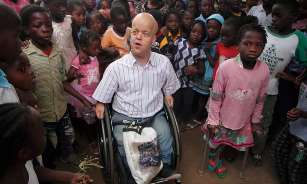 A man in a wheelchair looks to his right. He is surrounded by school children. They are in Mozambique.