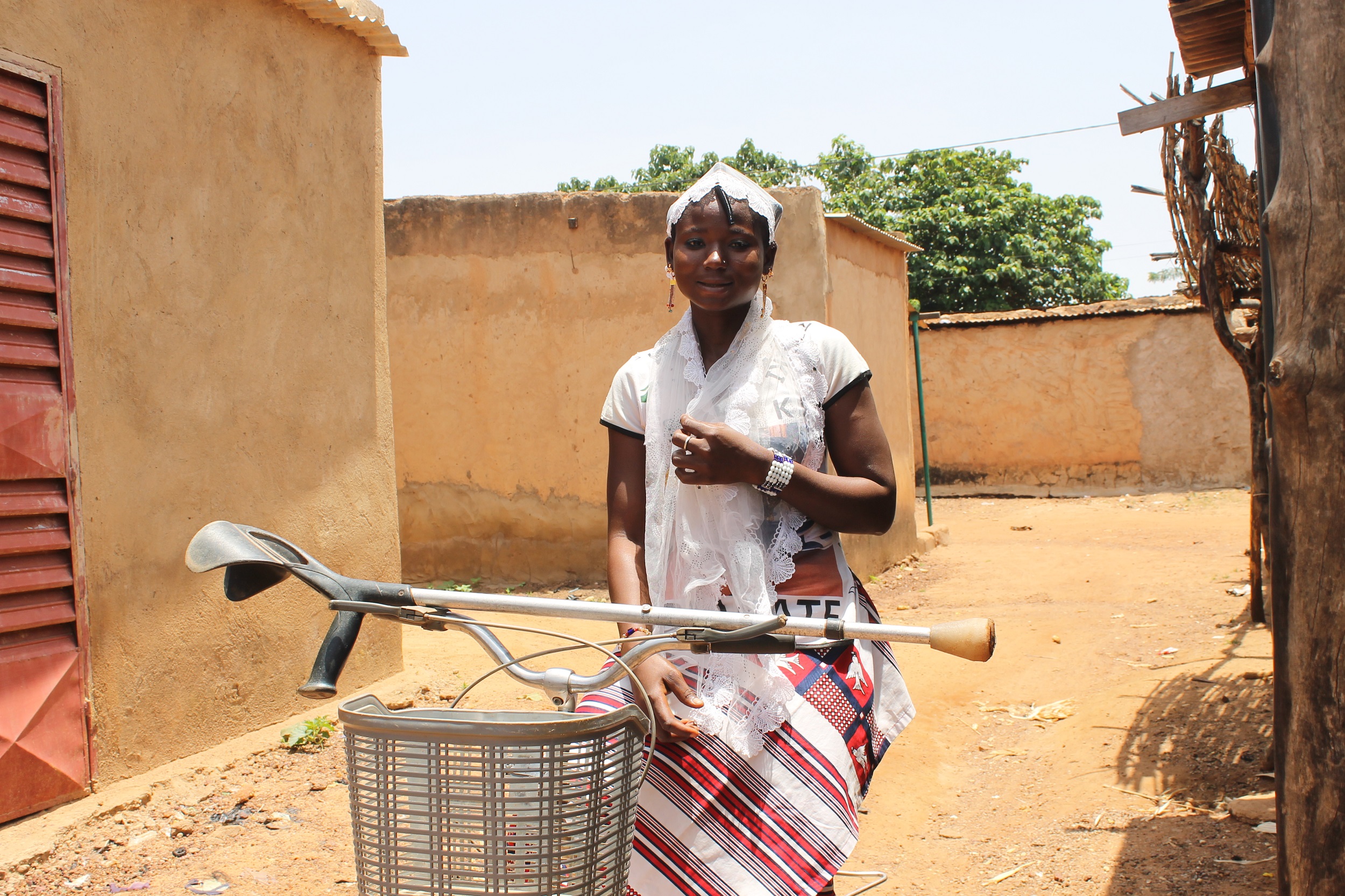 a young burkinabe woman on her bicycle, wearing a white lace head cover and matching shawl. Her skirt has red, white and black stripes. her crutch lies horizontally on the bike's handles.