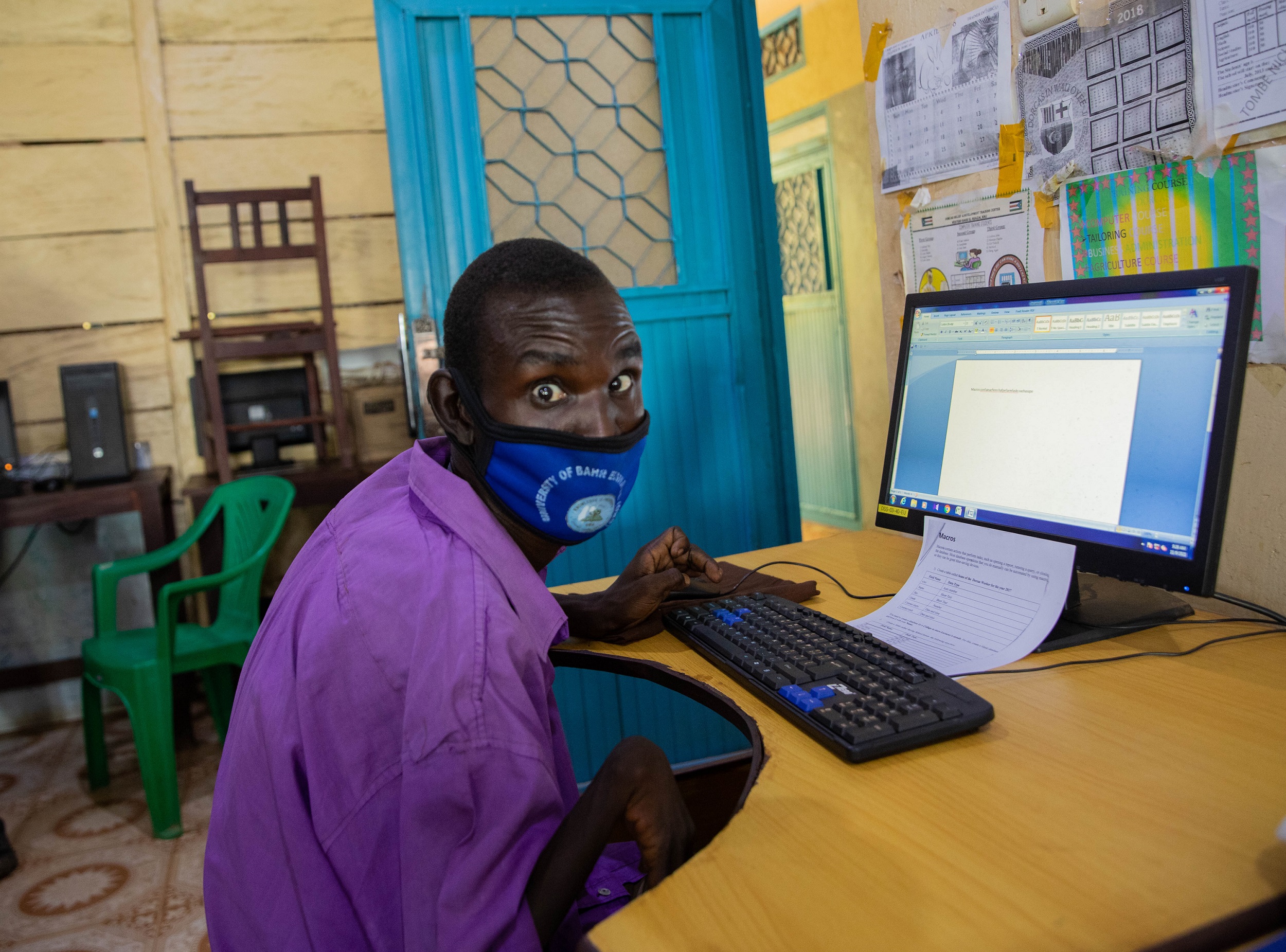 a young South Sudanese man wearing a blue facemask and lilac dress shirt sits at a desktop computer in a training centre.