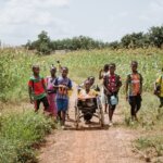 a young burkinabe girl in a wheelchair in the middle of her friends on a dirtpath through a cornfield