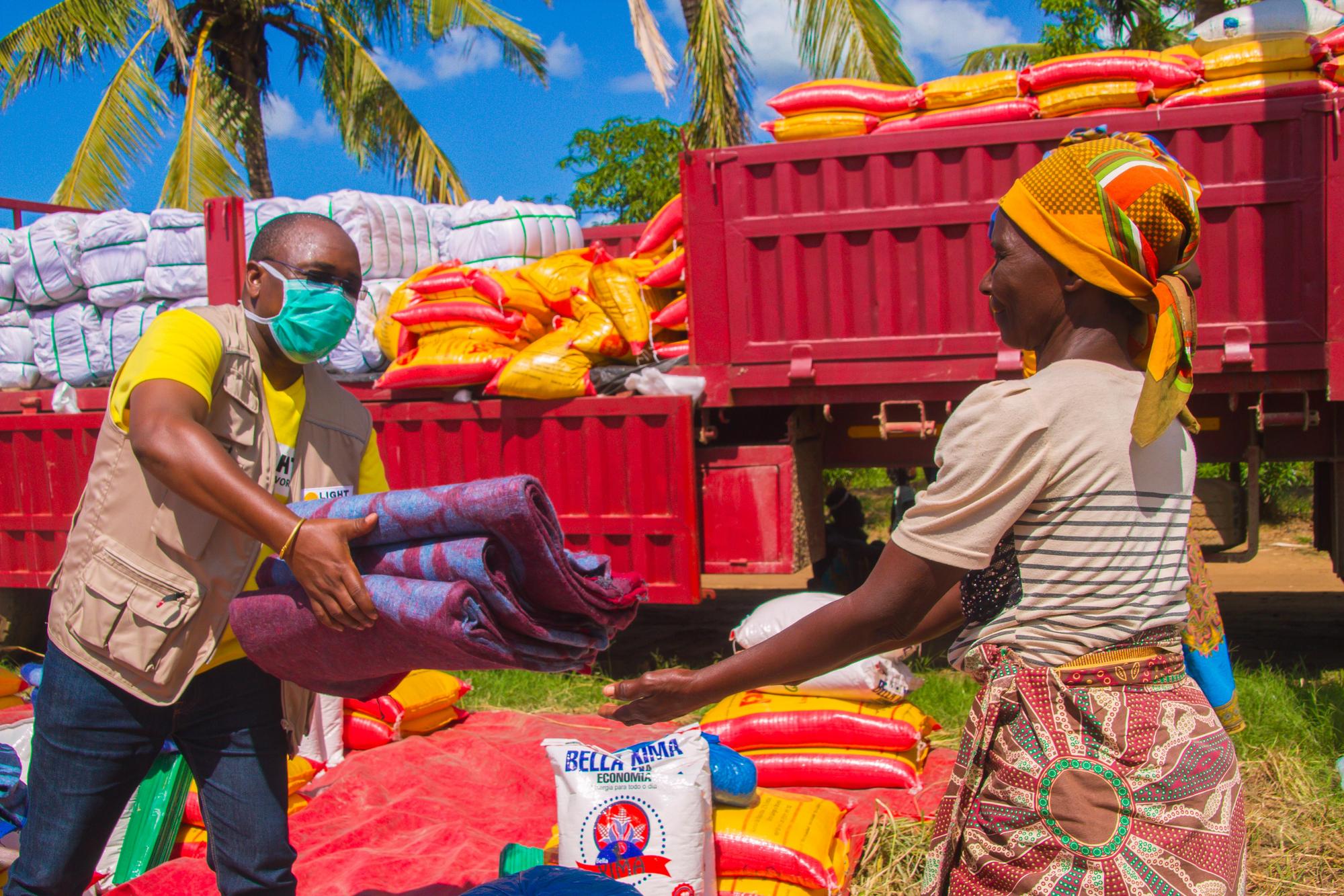 A slideshow image shows an aid worker from Light for the World wearing a yellow T-Shirt and a mask hands blankets to a woman from Mozambique dressed in a beige shirt and a colorful cloth on her head.