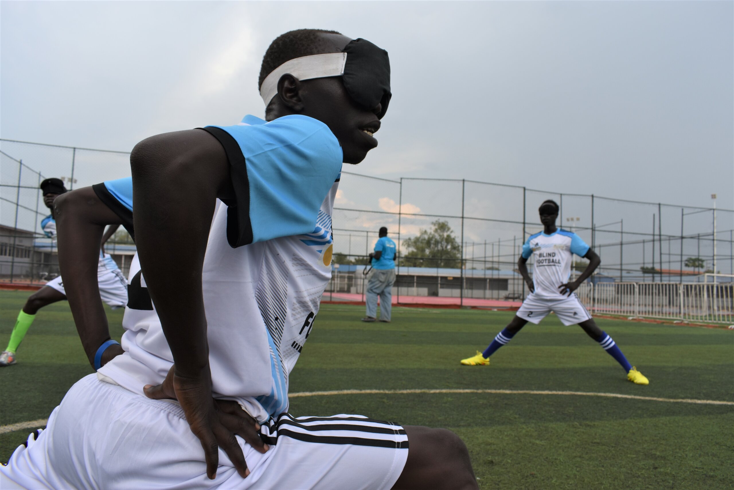 A young man from South Sudan is stretching in a soccer jersey with his teammates on a big field.