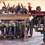 Hinter einer Holzschulbank sitzen viele Kinder und heben die Arme. Sie schauen nach rechts. Daneben sitzt ein Bub in einem Rollstuhl und hebt ebenfalls lachend die Arme.