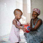 A young child with is playing with a red ball while her mother is kneeling next to her with a big smile. The girl is wearing glasses she received from Light for the World.