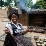 A woman with a black cloth on her head a white and black T-Shirt and a flowered skirt sits in her wheelchair and smiles into the camera. In the background you can see cakes in a form and a big oven.