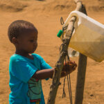 A young boy is washing his hand from a water canister that is fixated on wood.