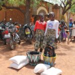 two Burkinabe girls wearing identical blue patterned skirts stand behind the food supplies and school kits they received