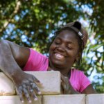 A woman in a pink shirt and a brown cap is working on a brick wall with a big smile in her face.