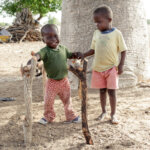 A boy with a disability practices walking on two wooden Bars. The child wears a green pullover and pink trousers. Next to it there is another boy supporting.