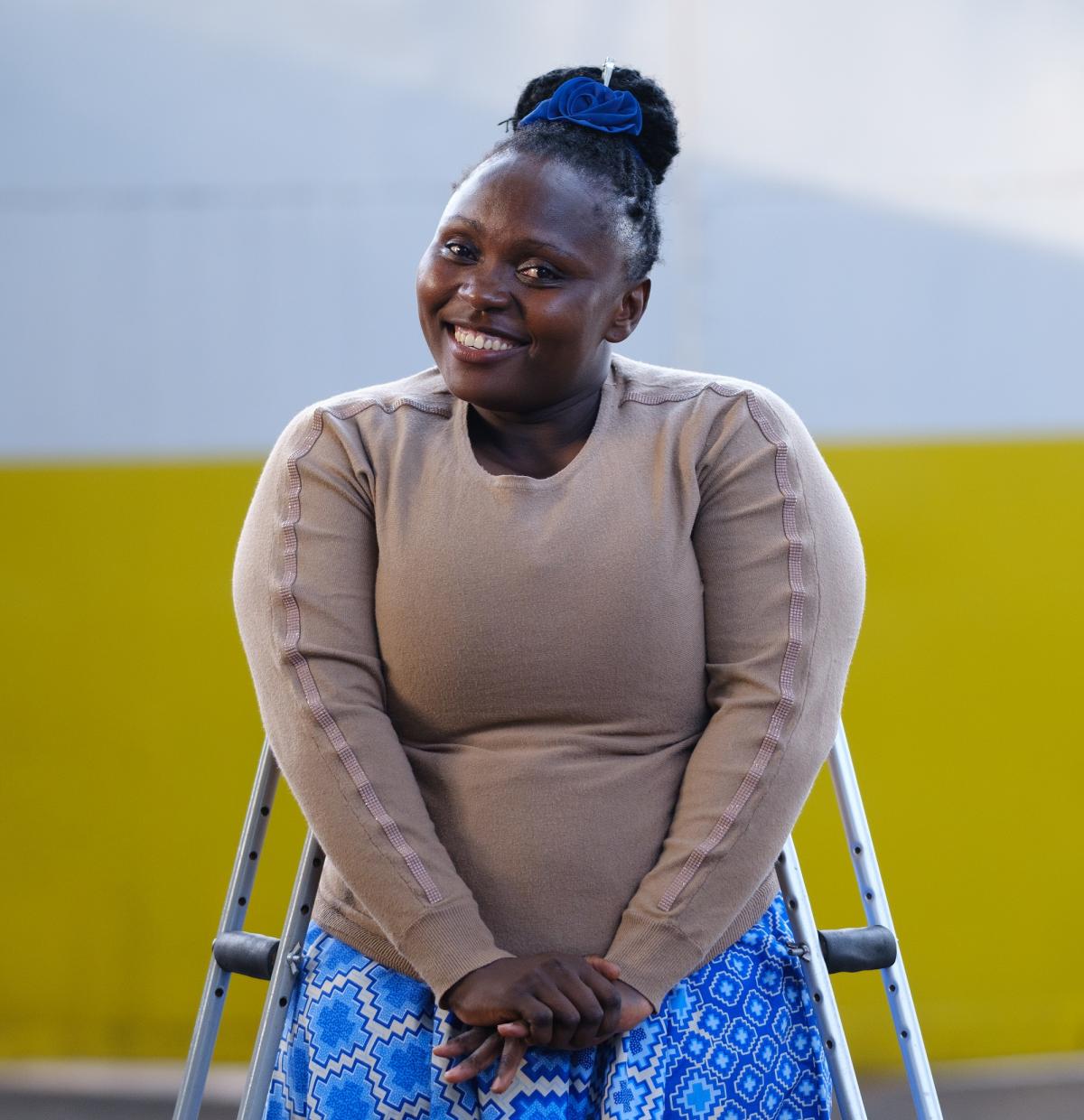 Dorcus Kabahenda, a 33-year-old Ugandan woman leaning on her crutches. she is smiling and wears a lightbrown sweater and a blue skirt