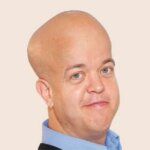 A headshot of Professor Tom Shakespeare. He is a white man, wearing a blue shirt in this picture. He has blue eyes. Likewise, he smiles at the camera. Tom has achondroplasia.
