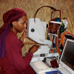 An ophthalmologist is conducting a eye exam on an elderly woman. The woman is looking into the machine while the ophthalmologist is holding the machine and looking at the computer screen.