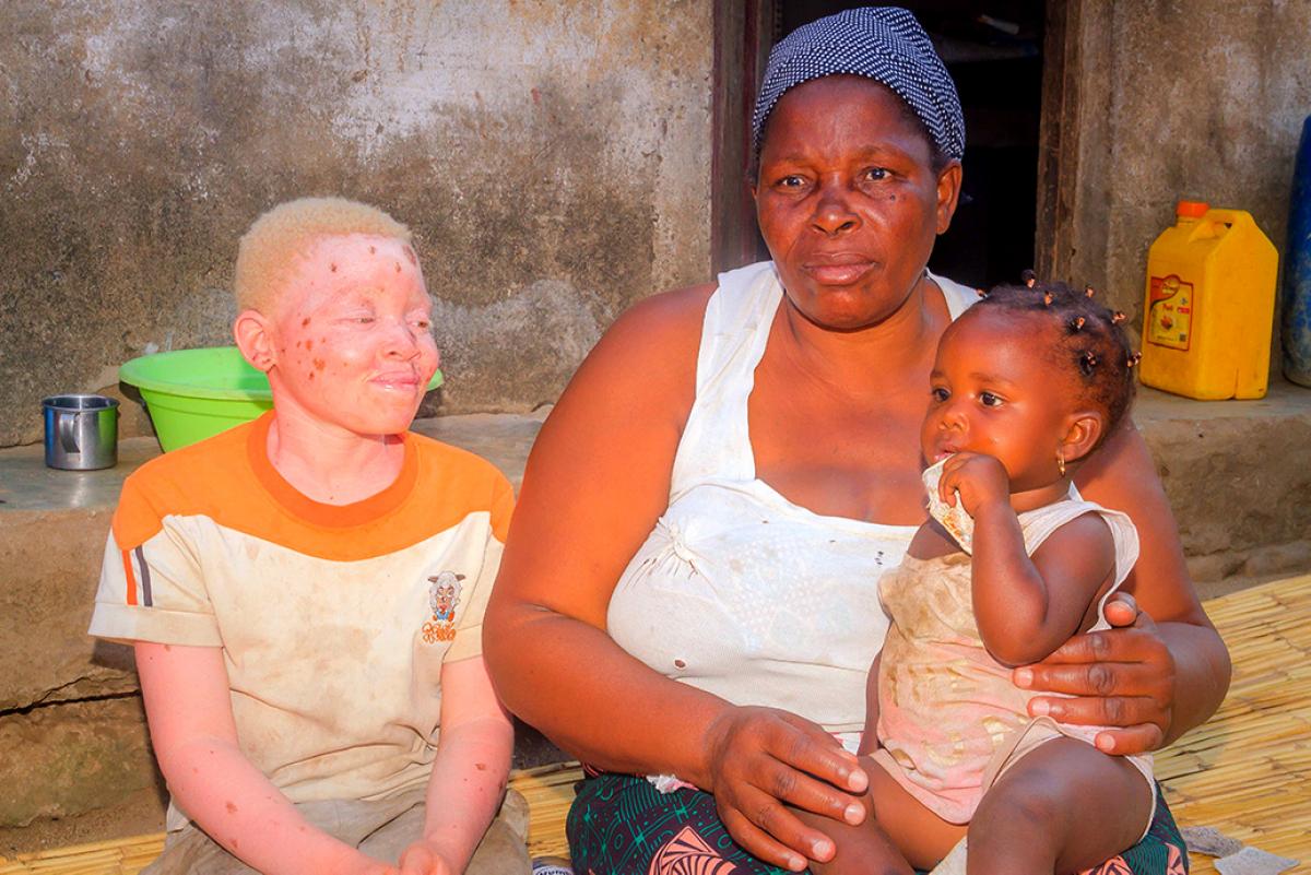 an older Mozambican woman with a blue and white patterned headscarf sits on a bamboo mat in front of her house. she holds her young granddaugther on her lap and her grandson with albinism sits next to her.