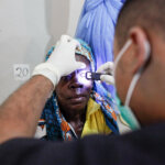 A woman from Mozambique with a colourful headscarf is being examined on her right eye. The man opposite her, wearing a face mask and white gloves, holds a bright light on her eye.