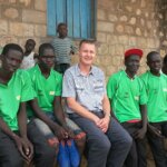 A European Man in a grey T-Shirt sits in between young meen wearing green T-Shirts.