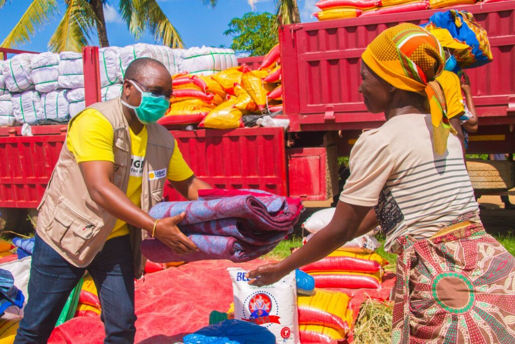 In front of a huge red lorry Light for World Mozambique Director Zacarias Zicai wearing a mask hands a blanket to a woman with a headscarf.