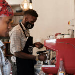 A young man with a protective mask and apron stands in front of a red coffee machine and prepares fresh coffee.