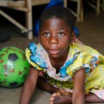 Child sitting on the ground next to a green football, leaning forward and looking up at the camera.