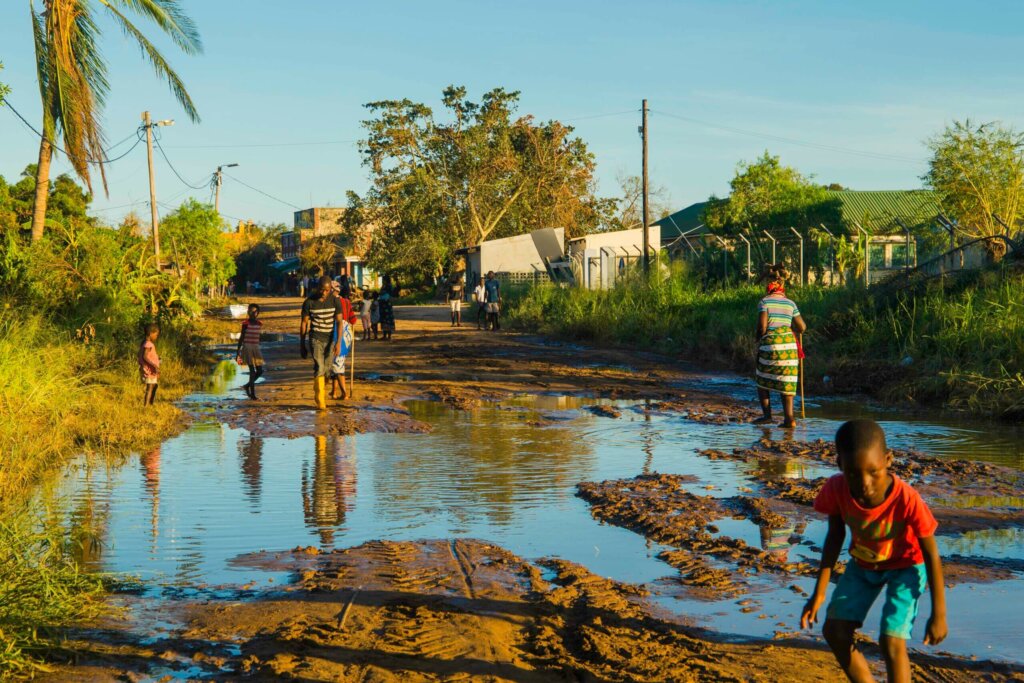 Image showing the flood that was left behind after cyclone Eloise. A child wearing a red top and blue shorts is standing on the right side of the image bending down whilst looking at the camera. In the back there are people walking across the piles of water.