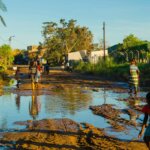 Image showing the flood that was left behind after cyclone Eloise. A child wearing a red top and blue shorts is standing on the right side of the image bending down whilst looking at the camera. In the back there are people walking across the piles of water.