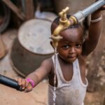 Small child looking at the camera while holding a water tank where water is coming out.