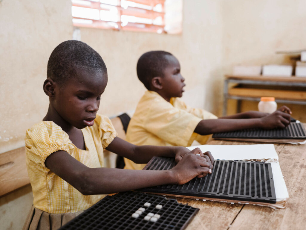 Two Burkinabe children in yellow school uniforms are sitting at a table learning Braille.