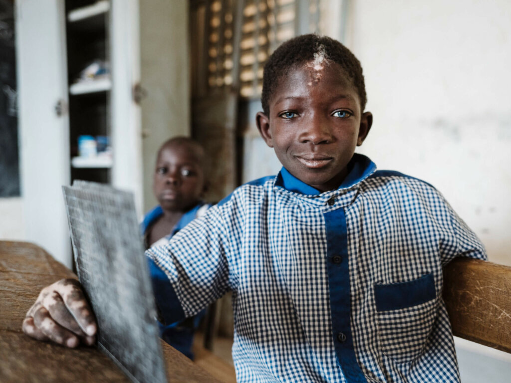 Vincent is a Burkinabe boy with a hearing disability. He sits in the classroom and looks directly into the camera with a knowing look.