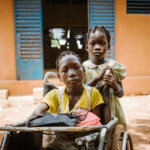 Two young girls are looking directly into the camera. One girl is sitting in a wheelchair whilst the other girl is standing right behind her holding the wheelchair.