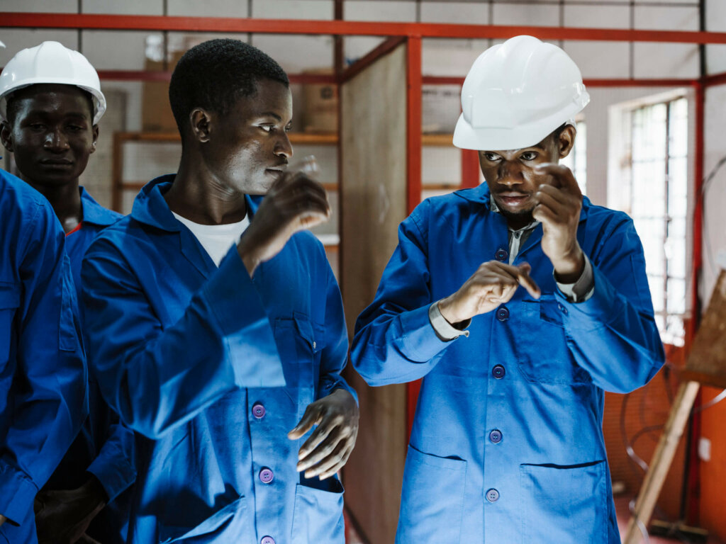 Three male Mozambican students in blue work overalls and hardhats communicate in sign language inside their training centre.