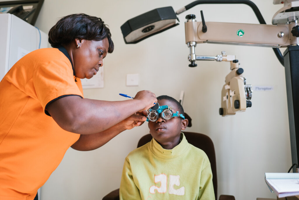 Ugandan boy Justine Basilica gets his eyes examined by a woman wearing an orange shirt. He is wearing exam spactacles to assess his visual acuity.